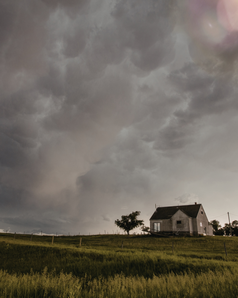 A small, weathered house sits alone on a grassy hillside beneath a dramatic sky filled with dark, heavy storm clouds, with a single tree nearby and soft light breaking through in the distance.