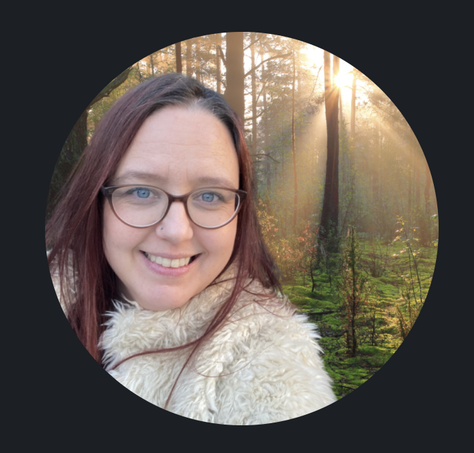 Helen Edgar smiling at the camera, with long auburn hair, glasses and a cream fluffy coat, standing in a woodland with sunlight streaming through tall trees and moss on the ground.