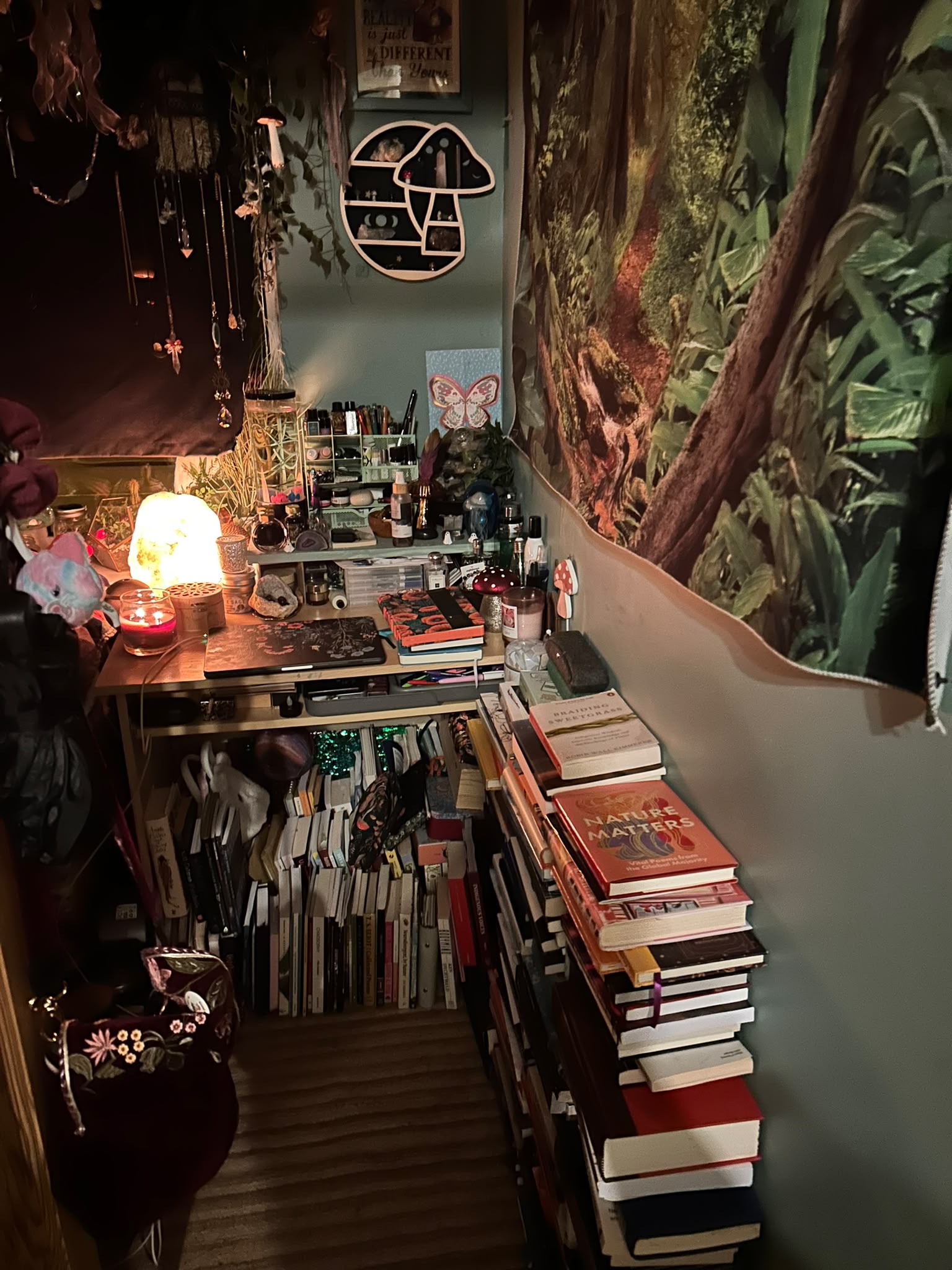A narrow corner of the den beneath a forest wall tapestry, with tall stacks of books including Braiding Sweetgrass and Nature Matters, shelves of books under the desk, and a mushroom-shaped wall shelf above.