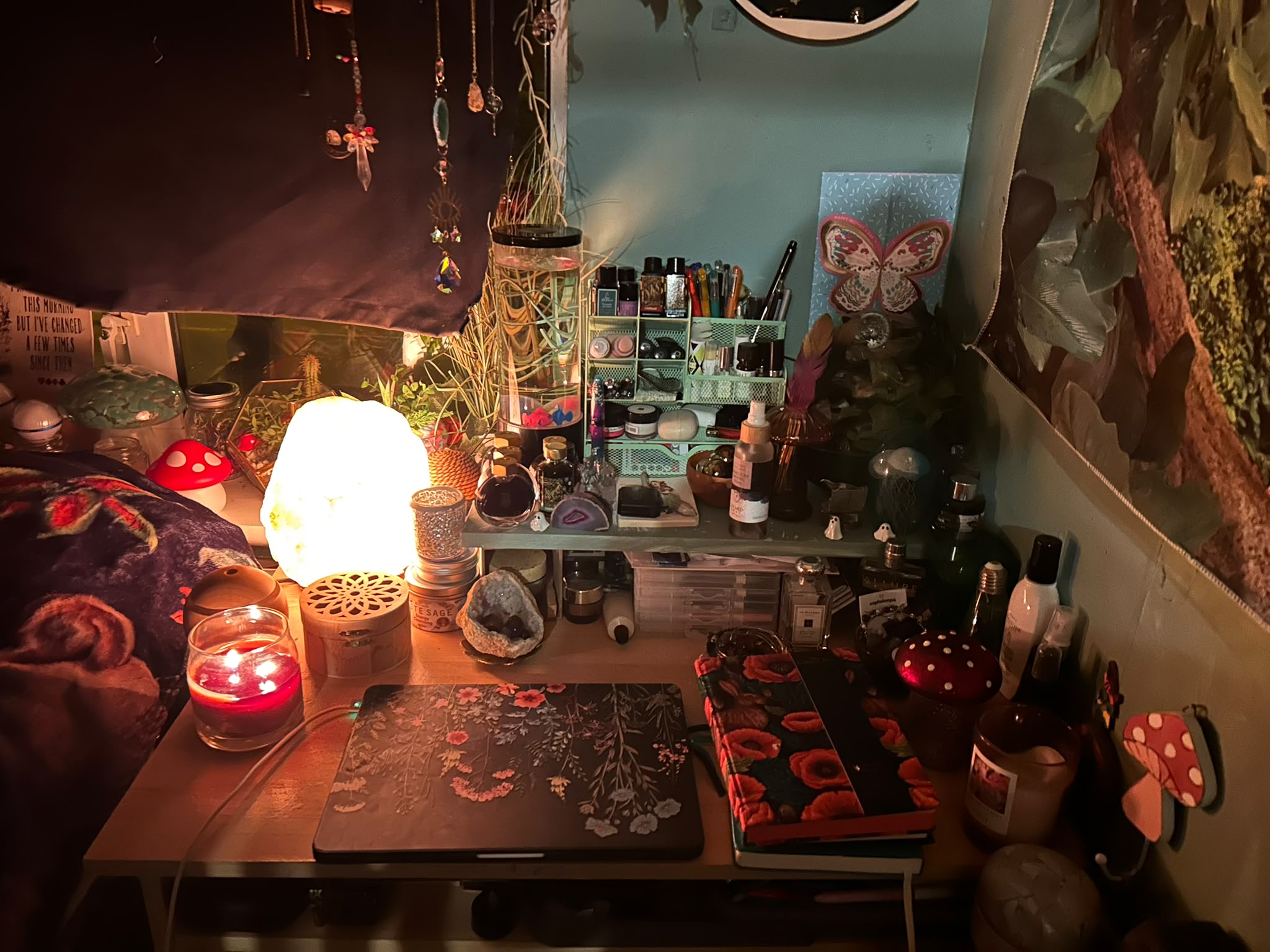 A writing desk in a dimly lit den, glowing with a salt lamp and a lit red candle. A laptop with a wildflower case and a poppy-covered notebook sit among inks, pens, crystals, mushroom ornaments and plants, with crystal suncatchers hanging above.