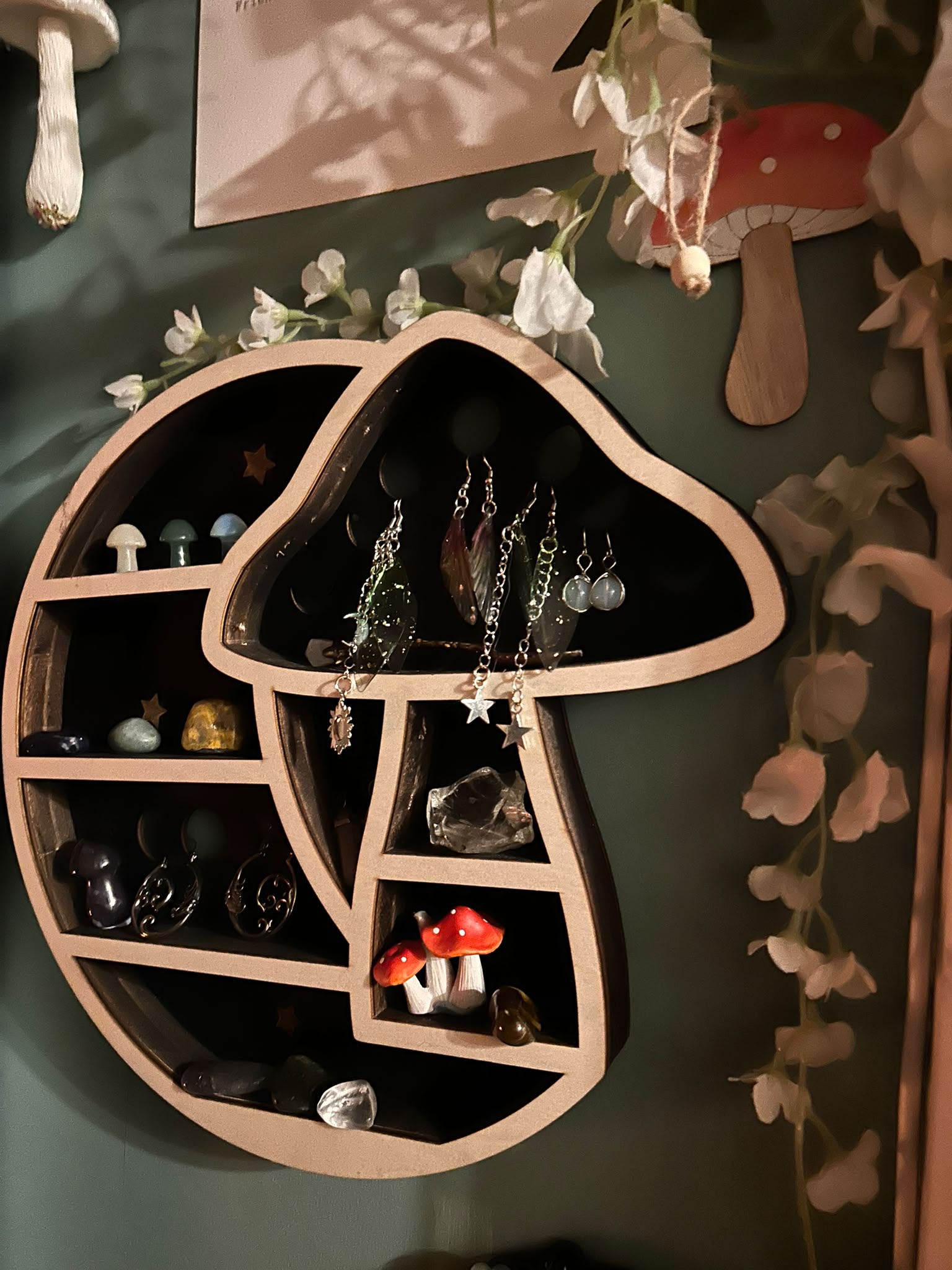 A wooden wall shelf shaped like a crescent moon and a mushroom, holding small crystal mushrooms, tumbled stones, faery-wing earrings and red-spotted toadstools, framed by trailing white flowers.