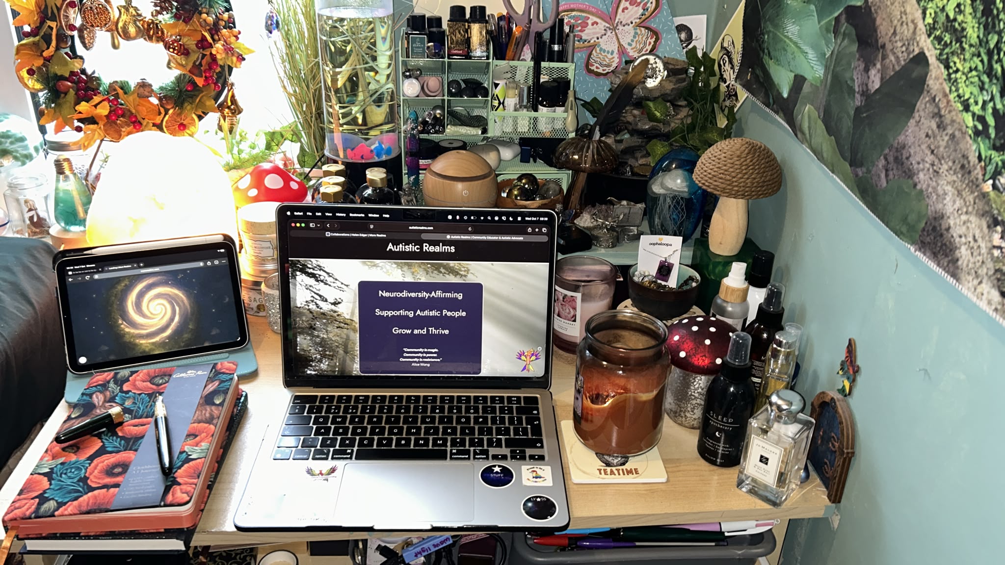 The writing desk by daylight. An open laptop shows the Autistic Realms home page, beside a tablet with a glowing spiral galaxy and a poppy-covered notebook with a fountain pen. Around them sit an autumn wreath, a salt lamp, a jellyfish lamp, mushroom ornaments, inks, candles and perfume bottles, with a forest tapestry on the wall.