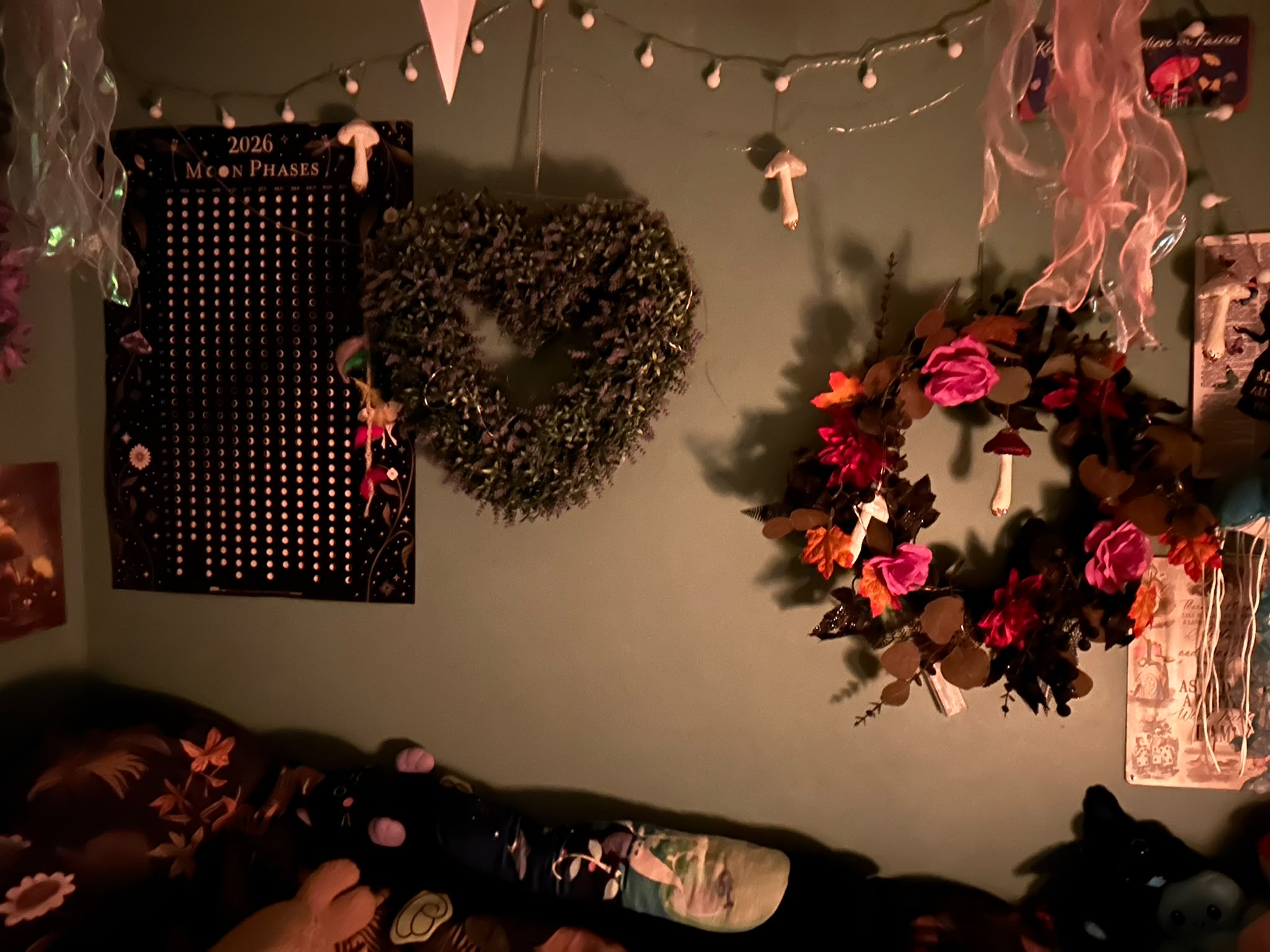 A sage-green wall with a 2026 moon phases poster, a heart-shaped lavender wreath and an autumn wreath of pink roses and orange leaves, under a string of small white lights and hanging white mushrooms.