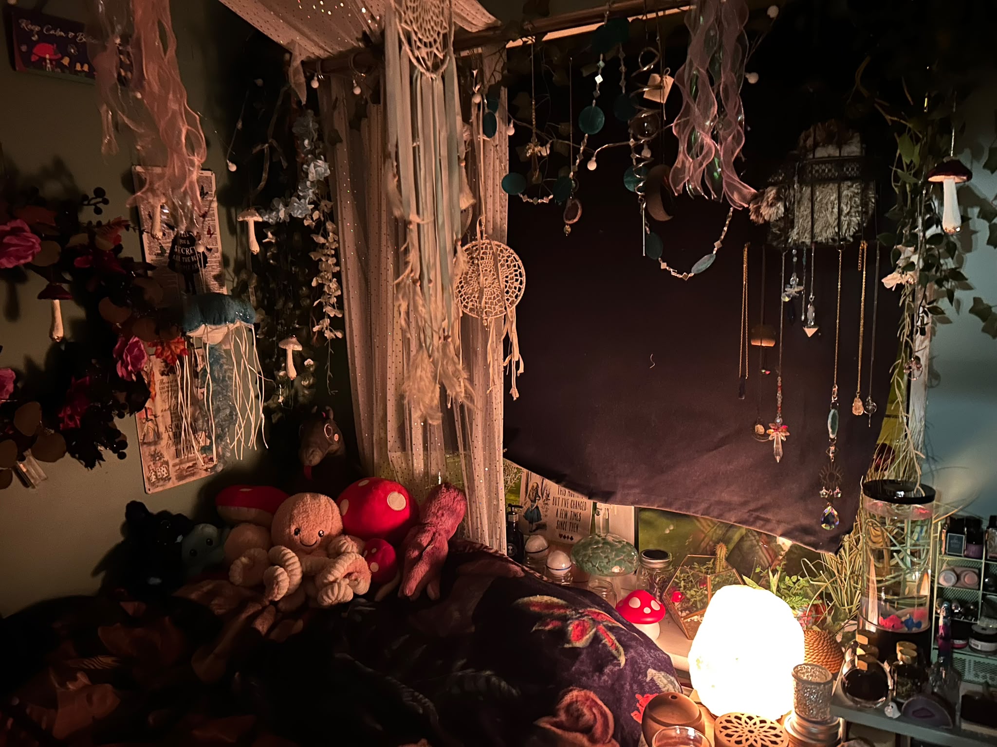 A cosy bed nook by a window draped with sheer curtains, dreamcatchers, ivy, ribbon jellyfish and hanging crystals. Soft toys, including an octopus and red-spotted mushrooms, rest on a floral throw beside a glowing salt lamp.