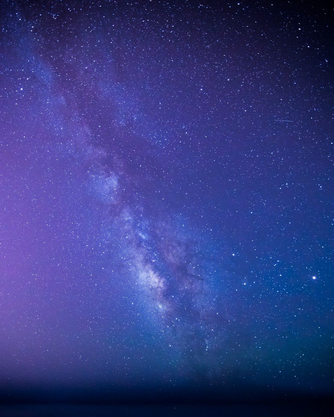 The Milky Way stretching diagonally across a lilac and deep blue night sky full of stars, above a dark horizon.