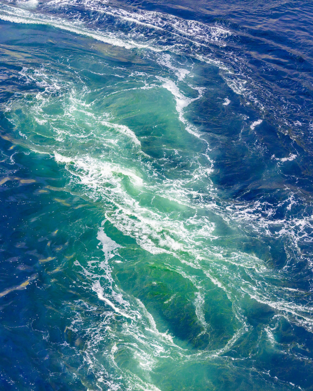 Aerial view of swirling ocean water. Bright turquoise-green currents spiral through deep blue sea, traced with curving lines of white foam, like a widening whirlpool.